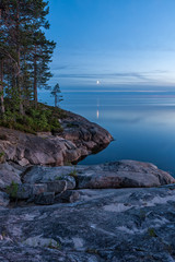 View on Onega Lake granite shore and evening glow with crescent reflecting in calm water at...