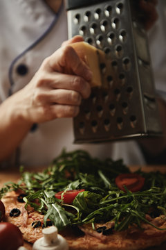 Italian chef grating parmesan, adding ingridients to pizza. European cuisine. 