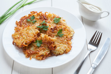 Fried potato pancakes or latke traditional homemade Hanukkah celebration food with greens and sour cream sauce recipe in plate on rustic white wooden table background. Natural organic meal