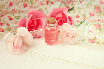Rose aroma extract fluid oil in cosmetic bottle, fresh garden rose flowers, floral pattern fabric. Feminine toiletries on shelf. Soft light and focus. Dreamy vintage pinkish tones edit.
