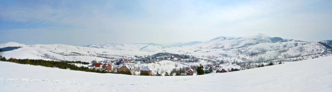 Panoramic image idyllic snowy winter landscape in the mountains, on a sunny morning, with snow-covered rooftops of a small picturesque village. Horizontal panorama.