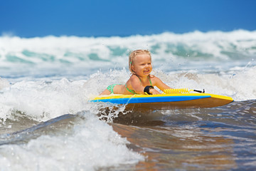 Fototapeta na wymiar Little baby girl - young surfer with bodyboard has a fun on small sea waves. Active family lifestyle, people outdoor water sport lessons and swimming activity on surf camp summer vacation with child.