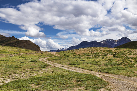 Winding dirt road among green meadows going to snow peaks