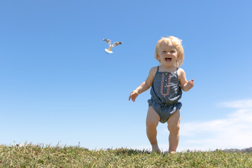 Happy blond baby girl runs on top of the hill on sunny summer weather, blue sky