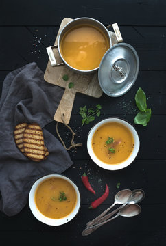 Red lentil soup with spices, herbs, bread in a rustic metal saucepan and bowls, over dark wood backdrop, top view