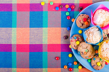 Fototapeta na wymiar Fresh homemade muffins and cookies with color chocolate coated candies on a colorful checkered kitchen towel. Free space for your text