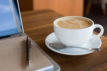 Hot latte art coffee cup on wooden table and note book on laptop