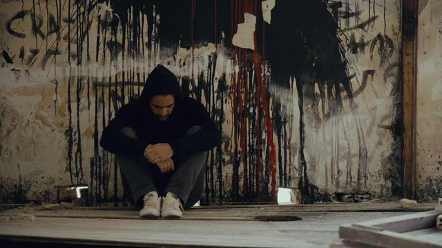 Hooded,troubled young man in wrecked abandoned building,back on graffiti wall.