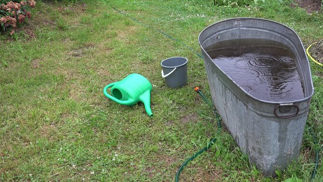 Tin water reservoir, watering can, bucket and water hose in rainy garden. Rain falling into water pool. Closeup shot. 4K
