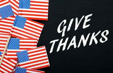 The phrase Give Thanks in white text on a blackboard next to miniature flags of the United States of America as a reminder for Thanksgiving