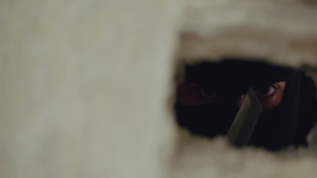 Panning shot of man in mask looking through a hole in the wall