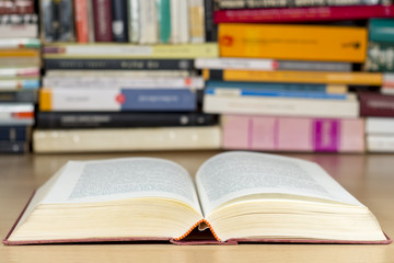 Open book on a wooden desk, with piles of books at background