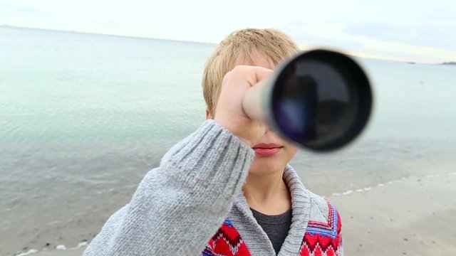 Funny little boy looking through telescope at camera at sea background. 8 years old child enjoying nature. Real time video clip.