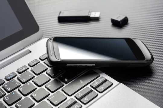 Blank Black Smartphone With Reflection Leaning On A Notebook Keyboard Next To An Open USB Storage Flash Drive, All Above A Carbon Layer
