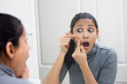 Closeup portrait of young frustrated woman surprised stunned to see zit on her face, gray turtleneck, isolated mirror reflection background. Facial feelings, situation, reaction