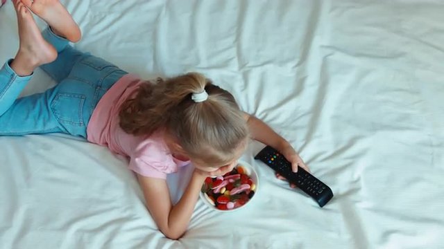 Girl watching TV lying on the bed. Child eating a sweets. Top view