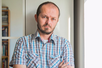 Fototapeta premium Man with beard in plaid shirt standing next to window, posing and looking to camera