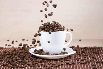 Roasted coffee beans falling down into white cup standing on white plate standing on tablemat on light wooden textured background.