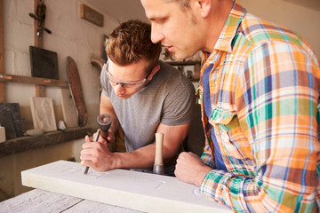 Stone Mason With Apprentice At Work On Carving In Studio