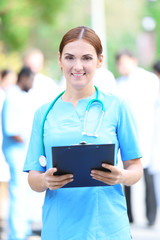 Beautiful woman doctor with clipboard in hands standing against unfocused group of medics, outdoors