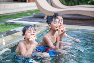 Three boys enjoying the pool while eating a bread