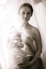 Portrait of beautiful young lady holding hands on pregnant stomach and covering with light fabric