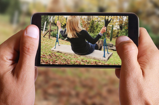 Male hand taking photo of Young woman sitting on swing in fall autumn park or garden. Happiness and freedom concept with cell, mobile phone.