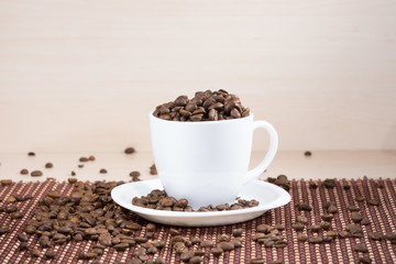 White cup full of roasted coffee beans standing on a white plate on tablemat on light wooden background.