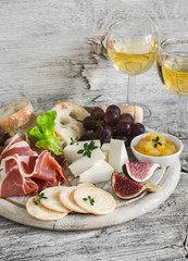 Ham, cheese, grapes, figs, nuts, bread ciabatta, cracker, jam on white wooden board and two glasses of white wine on bright wooden surface. Rustic style