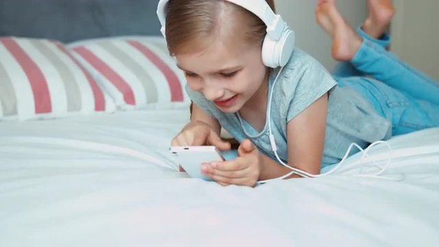 Portrait girl listening music in headphones and using smartphone cellphone. Child lying resting on the bed