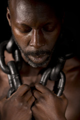 Black man with head bowed, holding large heavy chains around his neck.