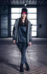 young woman with long hair, with a leather backpack, a warm hat, beautiful makeup, leather pants, a gray sweatshirt and a leather shirt, high heels walking one in the production hall and the street