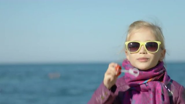 Portrait of a girl in sunglasses blowing bubbles on the sea beach. Child looking at camera