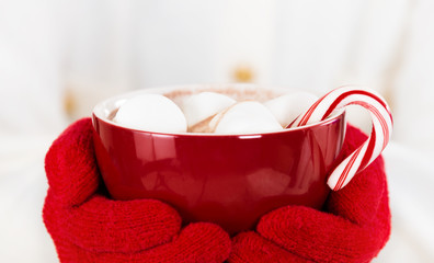 Mains gantées rouges tenant une tasse rouge de chocolat chaud avec des guimauves et une canne en bonbon