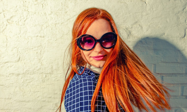 Trendy carrot-top girl turn around with her hair fly about her. Young woman weared in 60th fashion style sunglasses look back. A lot of copy space on white wall.