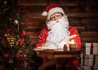 Obraz na płótnie Canvas Santa Claus in wooden home interior sitting behind table and writing letters with quill pen
