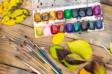 Plastic paint palette with paint, brushes and yellow leaves on w