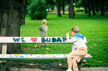 Little boy sitting on a bench, and another little boy walking on grass