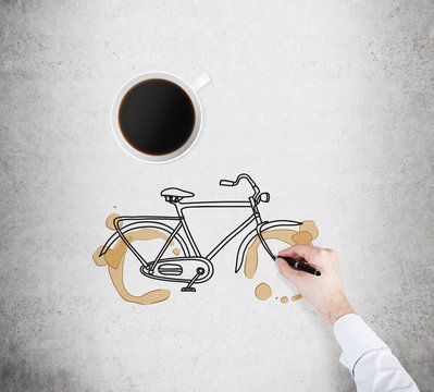 Fototapeta Top view of a cup of coffee and a drawing process of a bicycle on the concrete surface. A hand in formal white shirt with a pen.