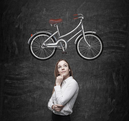 Fototapeta na wymiar A beautiful girl in formal clothes is dreaming about a new bicycle. A sketch of a bicycle is drawn on the black chalkboard.