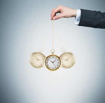 A hand holds a gold pocket watch in a chain as a pendulum. Light blue background. Time is money concept.