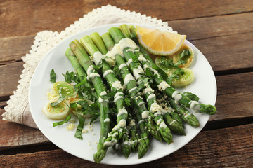 Appetizing asparagus in sour cream sauce with onion, cheese and lemon on white plate against wooden background