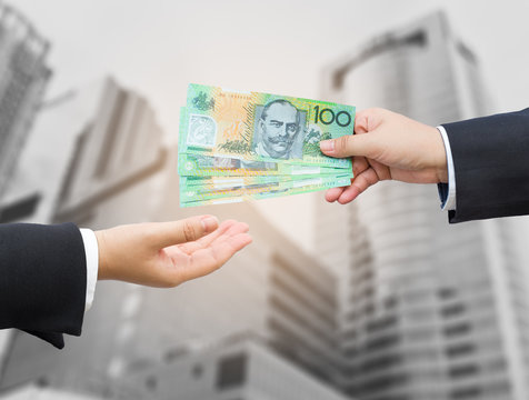 Hands of businessman passing Australian dollar (AUD) banknote with office building background.