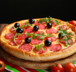 Tasty pizza with salami and olives decorated with tomatoes on wooden table against black background