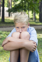 Sad boy hugging her knees and sits on green grass in nature.