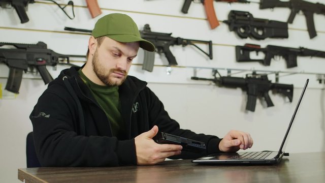 A man working at a laptop in gun shop, holding a gun in his hand