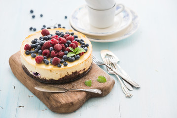 Cheesecake with fresh raspberries and blueberries on a wooden serving board, plates, cups, kitchen napkin, silverware over blue background