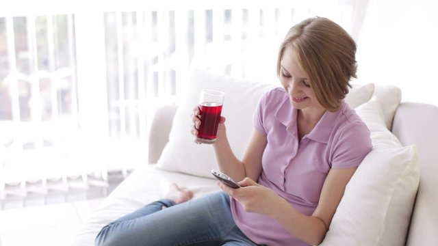Cheerful girl relaxing on sofa using cellphone drinking juice and smiling at camera. Panning camera