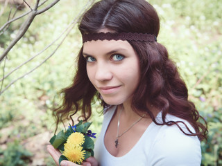 Naklejka premium Portrait of beautiful Caucasian girl with long red hair in white top with brown hippie style headband, with yellow flowers dandelions in forest park outside, summer spring day looking in camera
