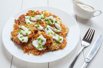 White plate of potato pancakes or latke traditional homemade vegan food with greens and sour cream on white vintage wooden table background